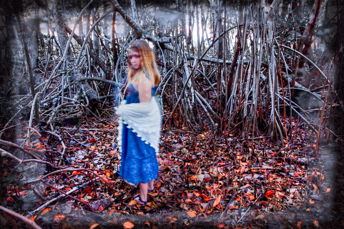 Figure in a blue dress and white wrap standing before dense mangrove roots
